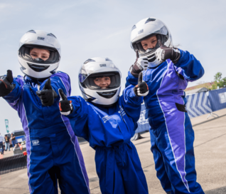 Drei junge Motorsportlerinnen sind in ihren Rennanzügen und schauen lächelnd in die Kamera. 