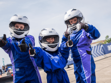 Drei junge Motorsportlerinnen sind in ihren Rennanzügen und schauen lächelnd in die Kamera. 