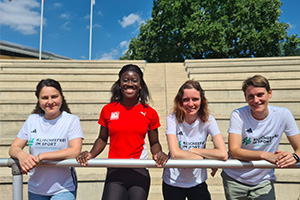 Die Sprinterin Yasmin Kwadwo-Bühner steht mit dem Projektteam von Klischeefrei im Sport vor einer leeren Tribüne.